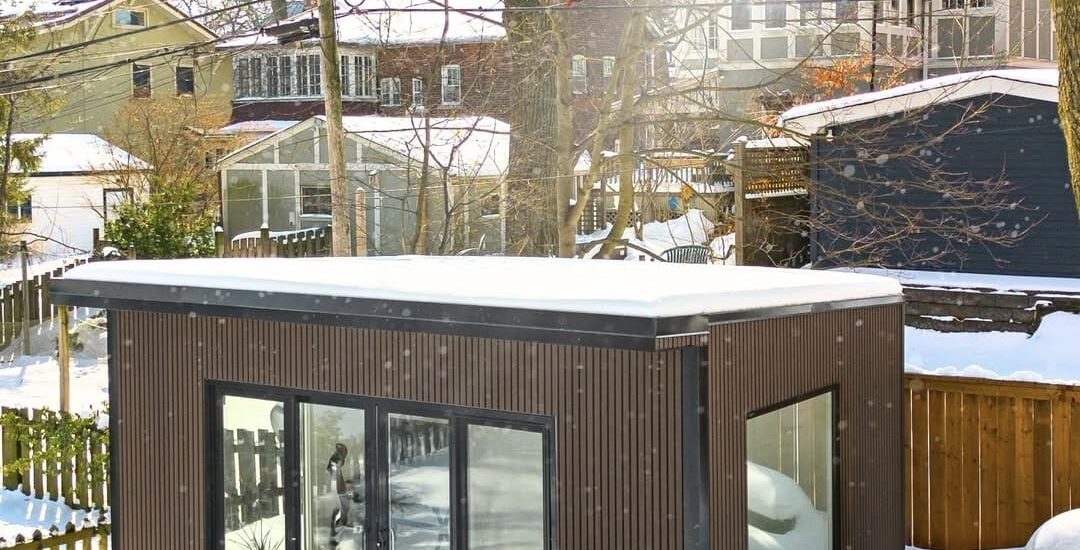 A modern, insulated garden room covered in snow, with warm interior lighting glowing through large windows, showcasing a cozy, winter-ready outdoor living space.