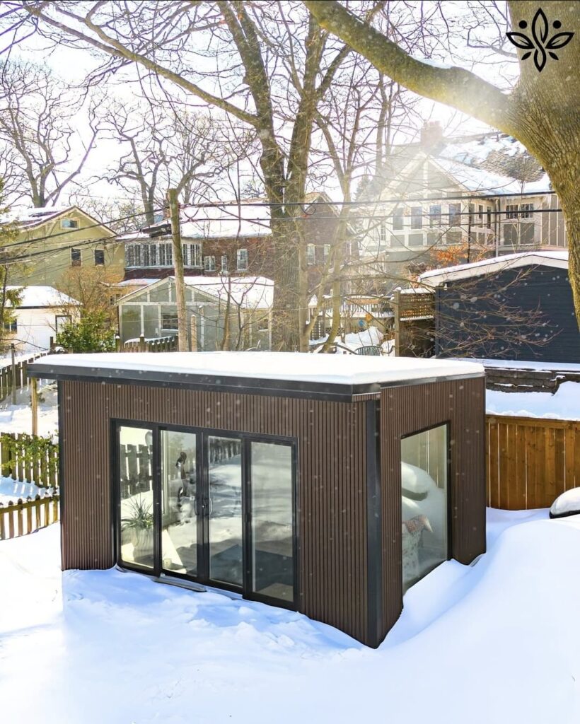 A modern, insulated garden room covered in snow, with warm interior lighting glowing through large windows, showcasing a cozy, winter-ready outdoor living space.