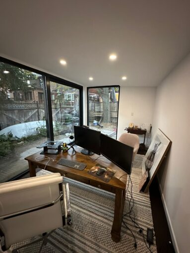 A beautifully designed garden room with a sloped roof, large windows, and a wooden deck, surrounded by lush greenery.