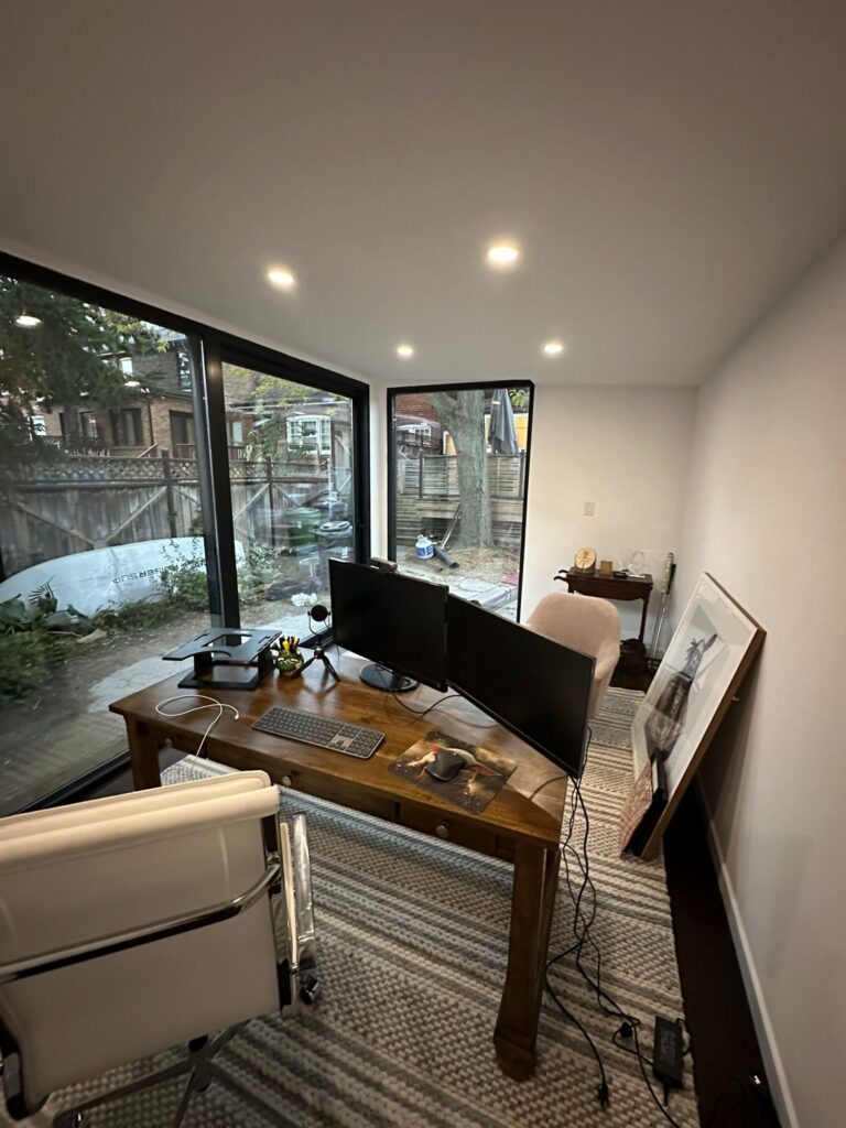 A beautifully designed garden room with a sloped roof, large windows, and a wooden deck, surrounded by lush greenery.