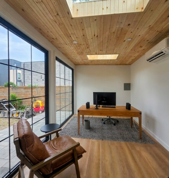 Modern custom garden room studio with floor-to-ceiling grid glass doors, dark wood fluted siding, and recessed soffit lighting on a stone paver patio.