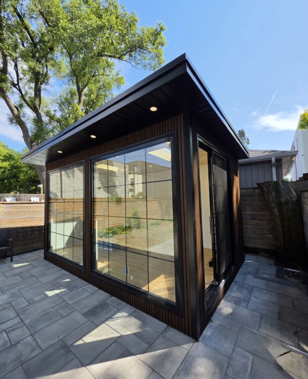 Modern custom garden room studio with floor-to-ceiling grid glass doors, dark wood fluted siding, and recessed soffit lighting on a stone paver patio.