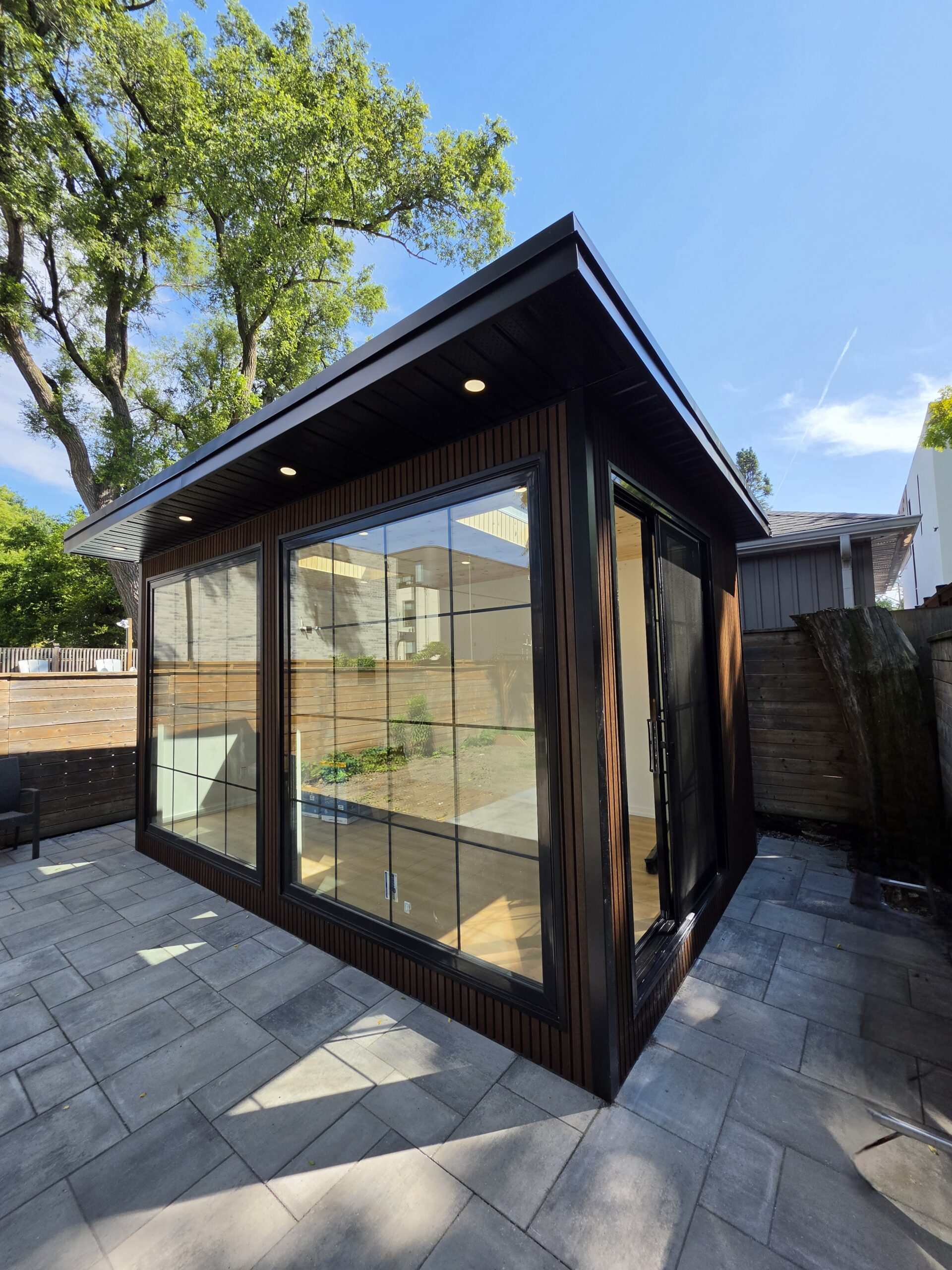 Modern custom garden room studio with floor-to-ceiling grid glass doors, dark wood fluted siding, and recessed soffit lighting on a stone paver patio.